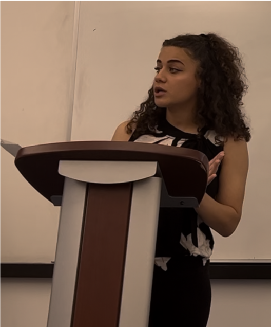 Young woman with curly hair speaking at a podium in a classroom or lecture hall.