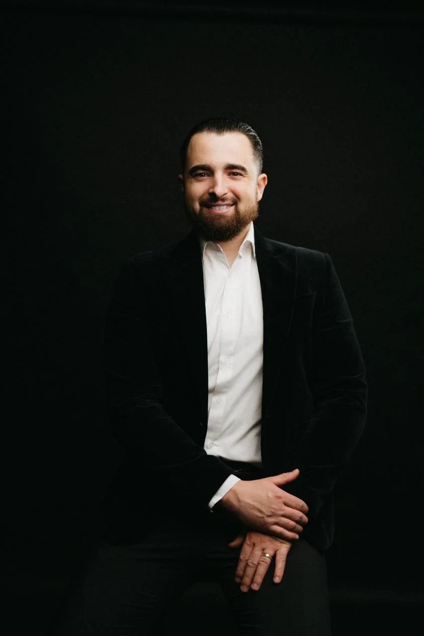 Un homme souriant, avec une barbe, portant un costume noir et une chemise blanche, pose contre un fond noir.