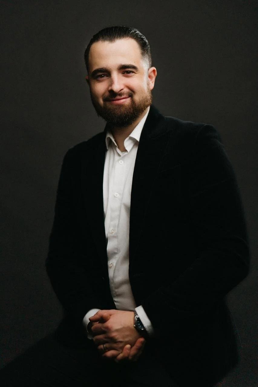 Un homme souriant, portant un costume noir et une chemise blanche, posé contre un fond noir.