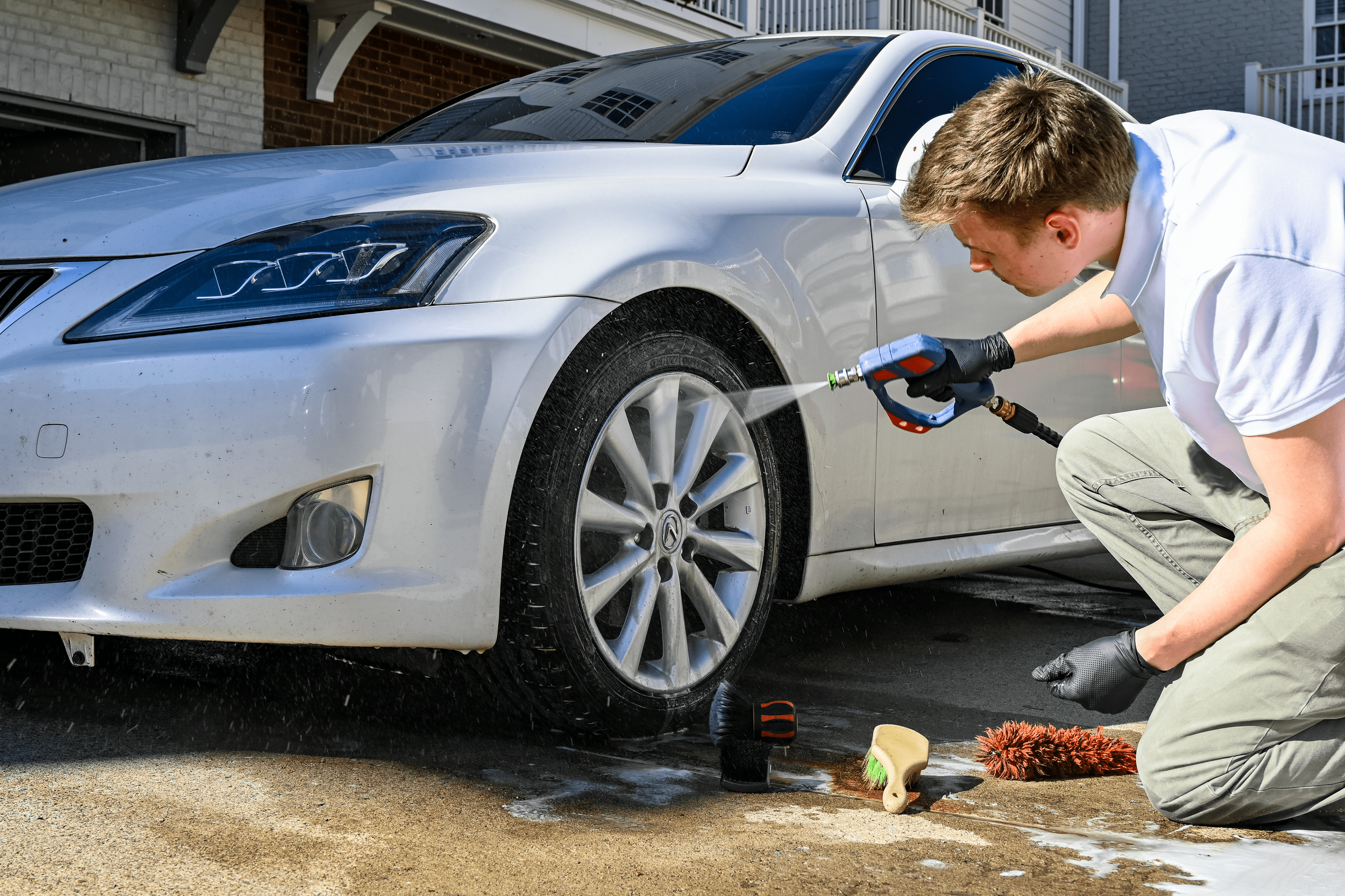 Person washing a black sports car with soap and sponge outside a building with brick wall.