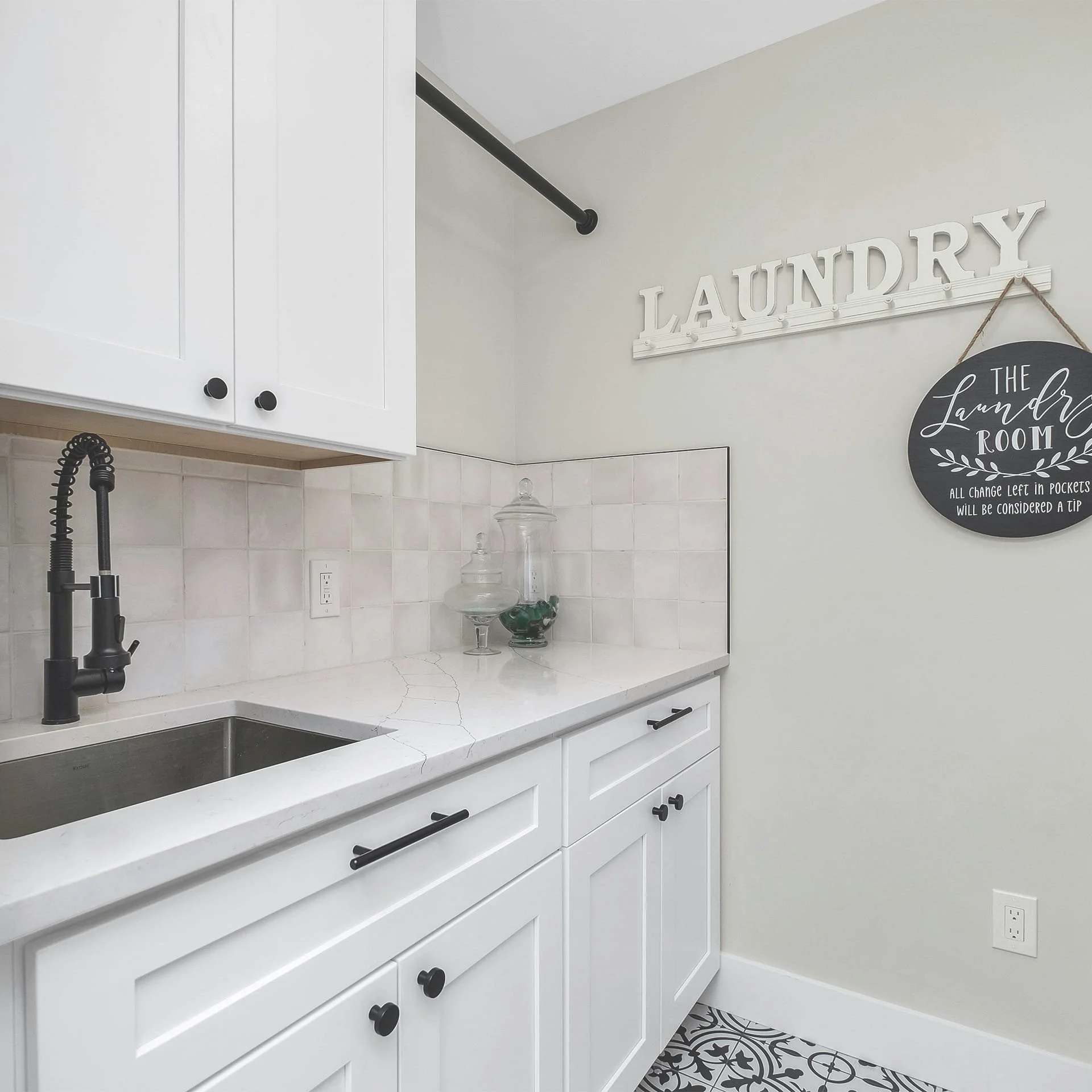 A laundry room with white cabinets, a black faucet, a white countertop with cracks, decorative glass jars, a sign that says 'LAUNDRY', and a decorative round sign that says 'The Laundry Room' with additional text.