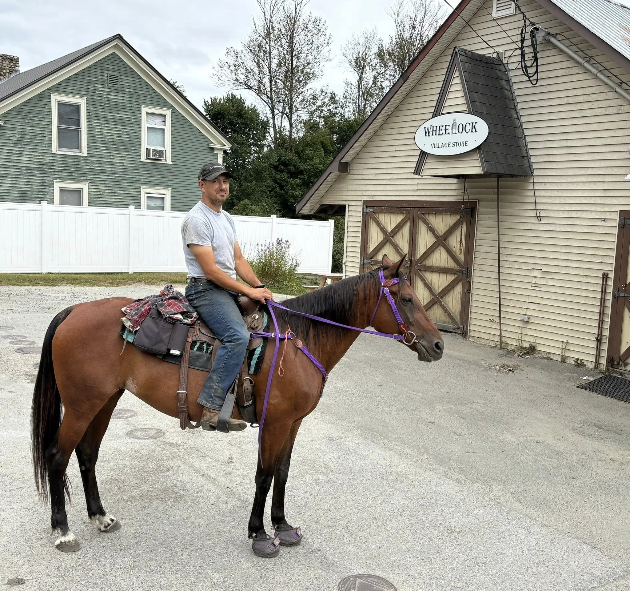 A man riding a brown horse with purple reins outside a village store named 'Wheelock Village Store'.