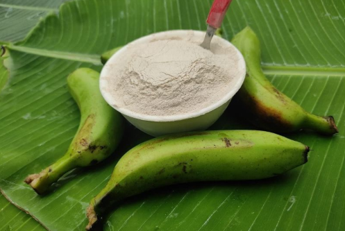 Three green bananas and a bowl of white flour on large green banana leaves.