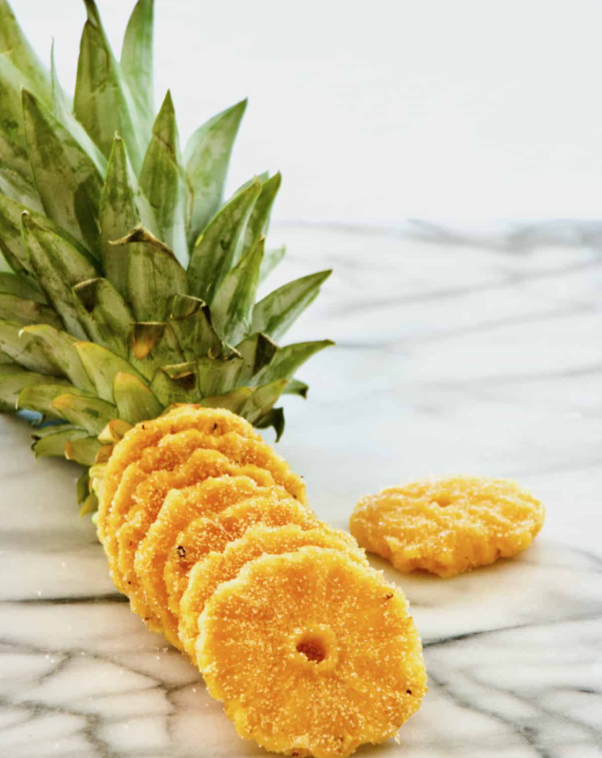Pineapple slices arranged on a marble surface with a peeled pineapple in the background.