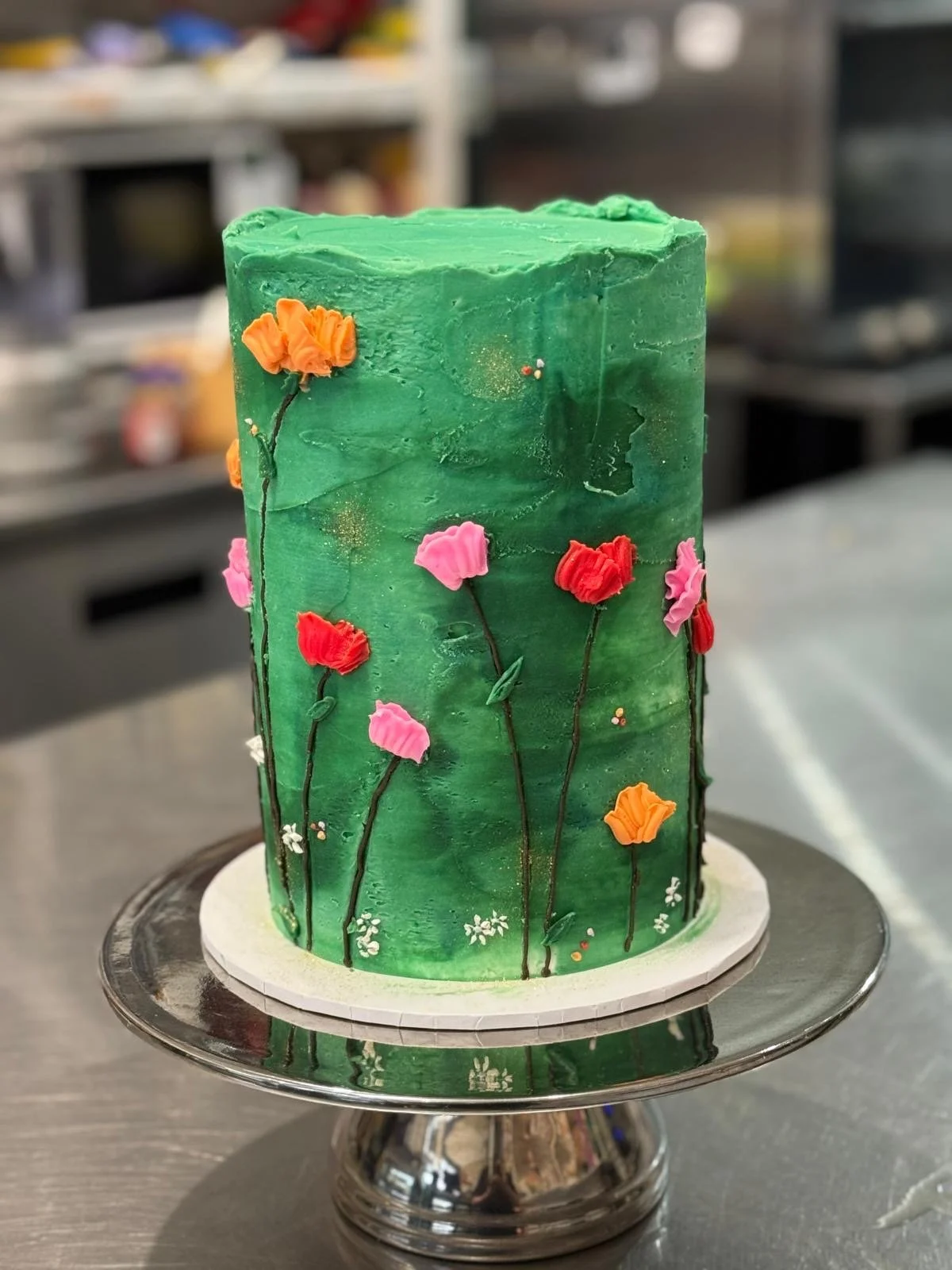 A tall, green, cylindrical cake decorated with colorful fondant flowers on thin black stems and leaves, resembling a garden scene.