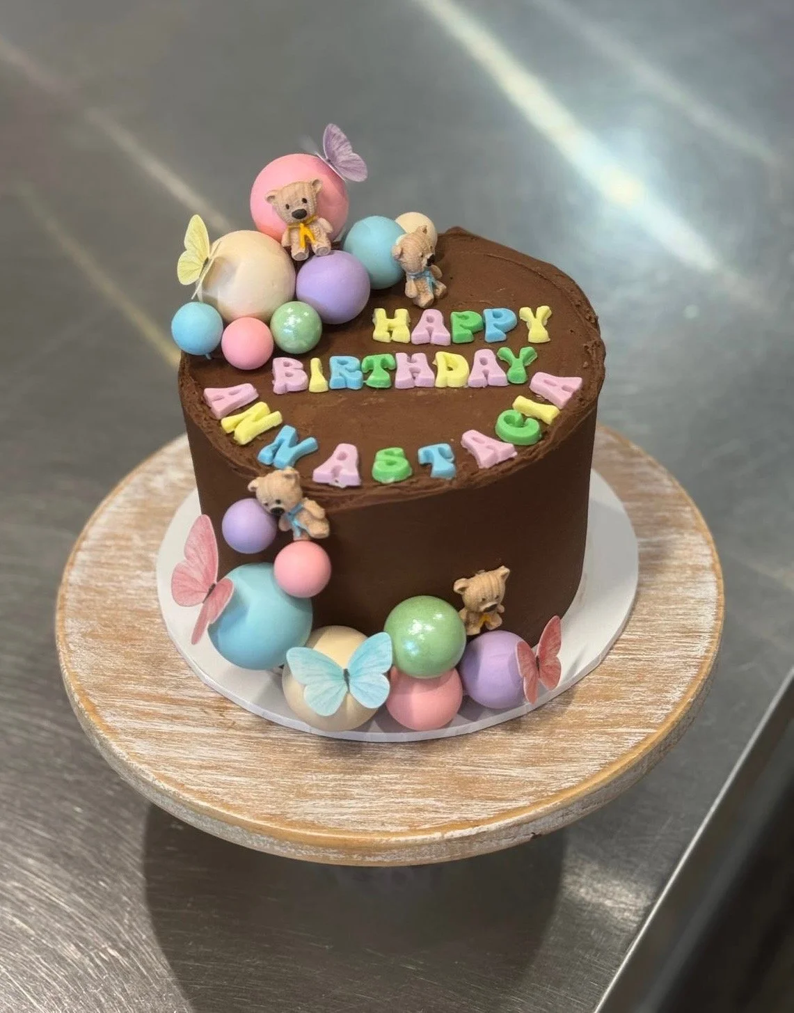 A chocolate birthday cake decorated with pastel-colored fondant letters spelling 'Happy Birthday' and 'Abaie,' surrounded by colorful fondant balls, pink and blue butterflies, and small teddy bear figurines, on a white cake board placed on a wooden s
