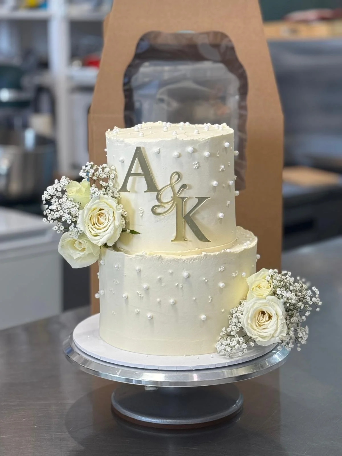 Two-tier ivory wedding cake decorated with white flowers and pearls, with gold initials A & K on top, placed on a silver cake stand.