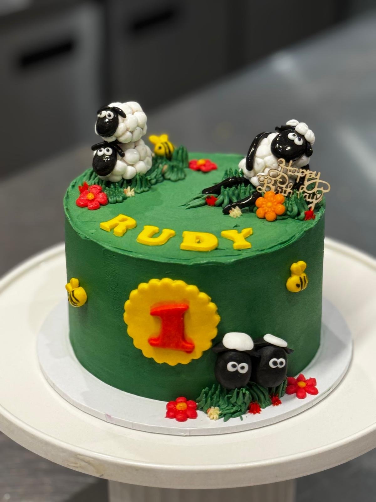 A green birthday cake with animal figurines, flowers, a yellow and red number one, and yellow letters spelling 'TRUDY'.