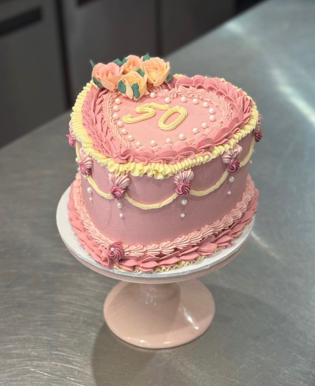 Pink heart-shaped cake with yellow and pink floral decorations on top, decorated with pink and yellow icing along the edges, and the number 50 written on top. The cake is placed on a pink cake stand.