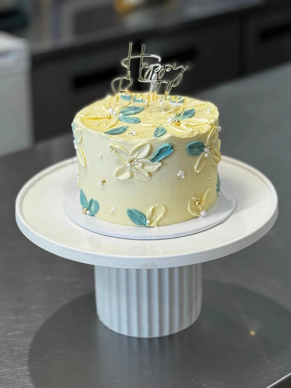A round cream-colored birthday cake decorated with blue and white flowers, small white and gold pearls, and a cake topper that reads "Happy Day" in metallic script, placed on a white pedestal cake stand.