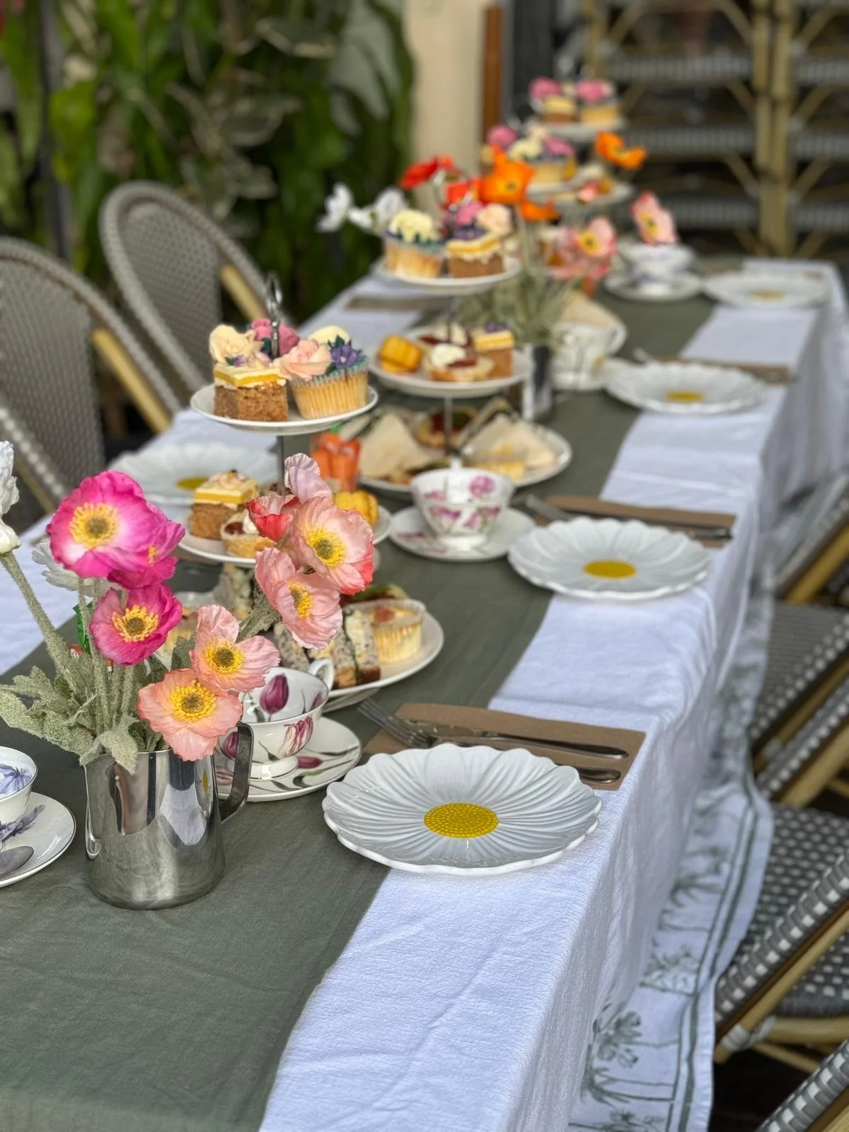 A long table set for afternoon tea with floral decorations, tiered trays filled with assorted cupcakes and desserts, plates, cups, and cutlery, with chairs around the table.