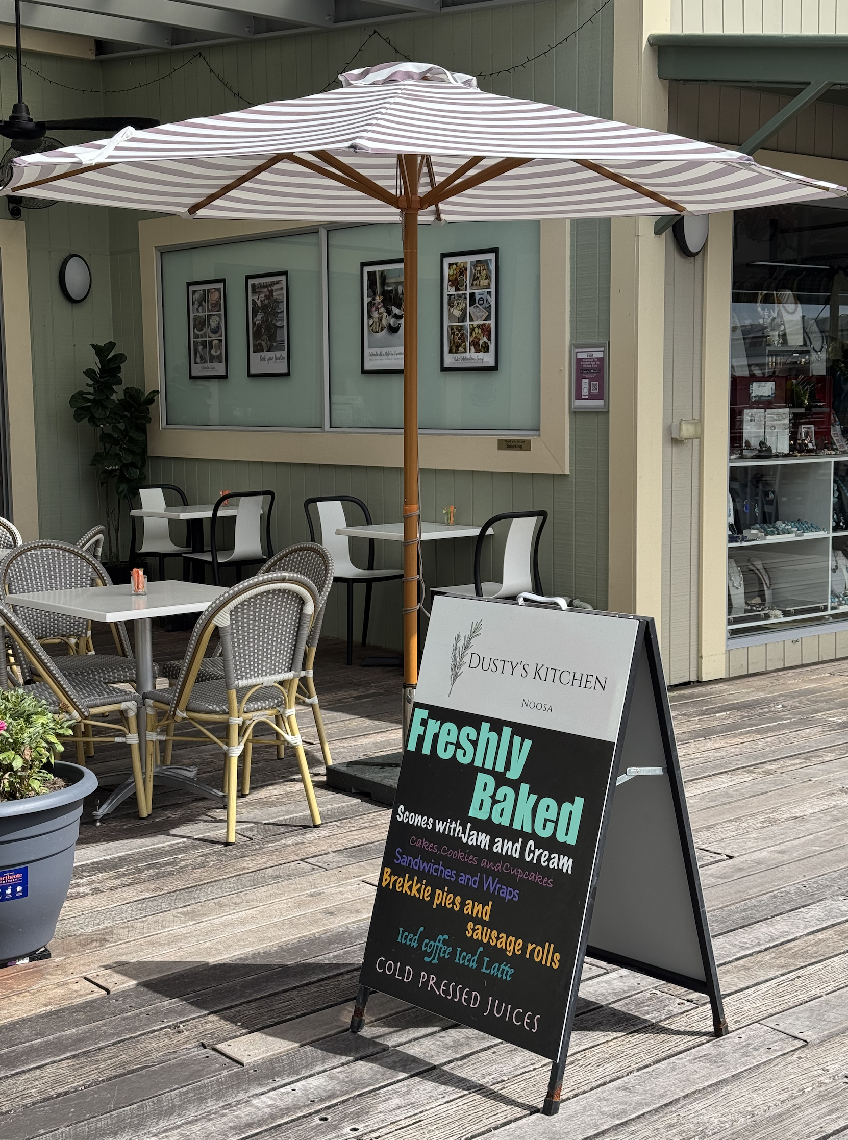 Outdoor patio with tables and chairs under a striped umbrella, a chalkboard sign advertising baked goods and breakfast items, and a storefront with framed pictures and a display of various products.