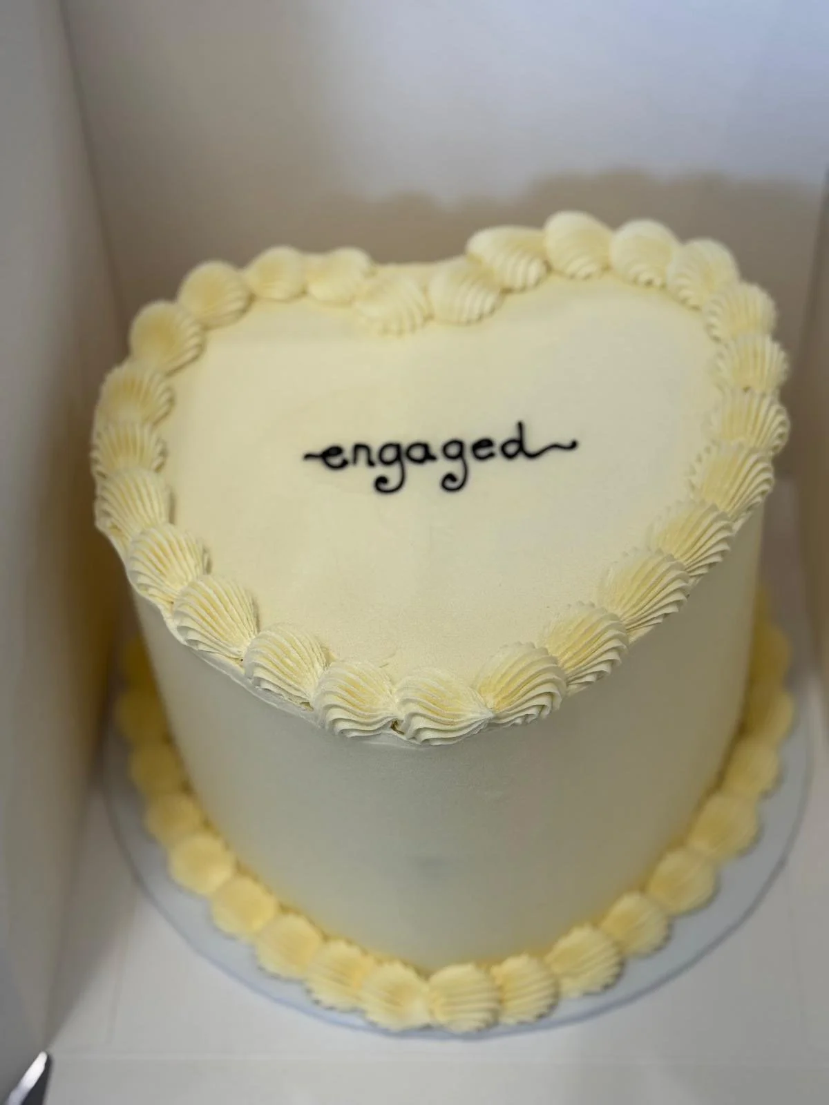 A heart-shaped white cake with yellow cream piping along the edges and the word 'engaged' written in black icing on top, inside a white box.