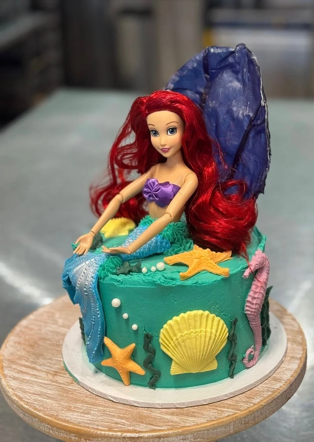 A mermaid-themed cake with a doll of Ariel sitting on top. The cake is decorated with starfish, seashells, seaweed, and an ocean wave made of fondant and icing.