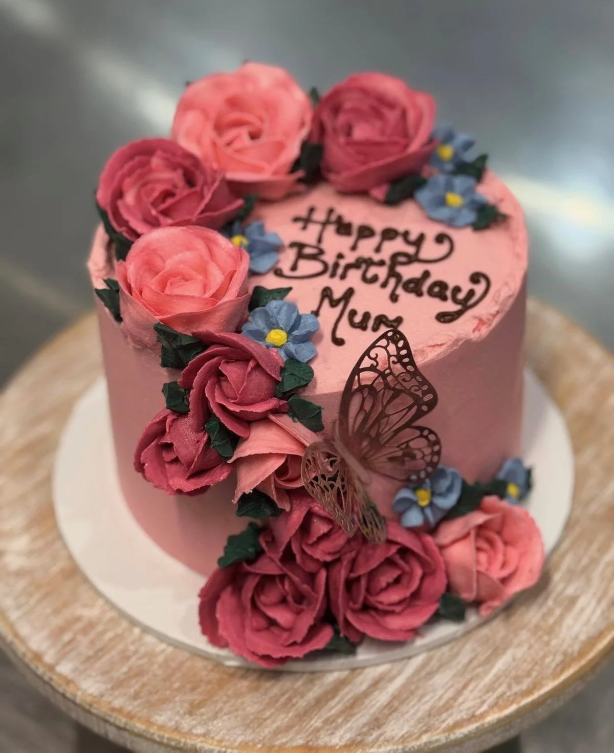 A round pink birthday cake decorated with pink rose-like frosting, blue flowers with yellow centers, and green leaves. The cake has a chocolate butterfly and a message written in black icing that says, "Happy Birthday Mum."