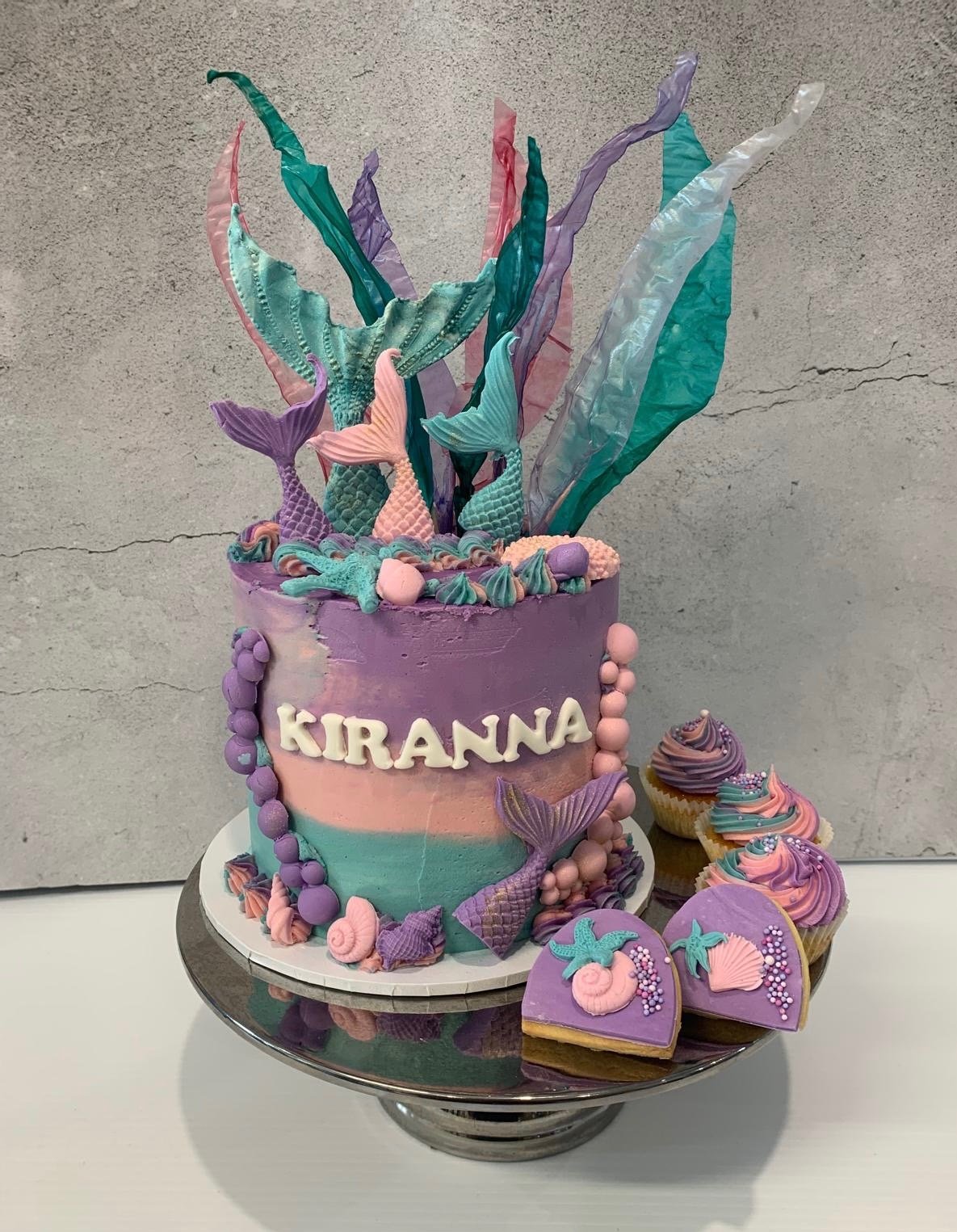 A colorful mermaid-themed birthday cake with pastel purple, pink, and green shades. The cake is decorated with mermaid tail and seaweed-shaped decorations, and the name 'KIRANNA' is written on the front. There are also matching cupcakes and cookies d