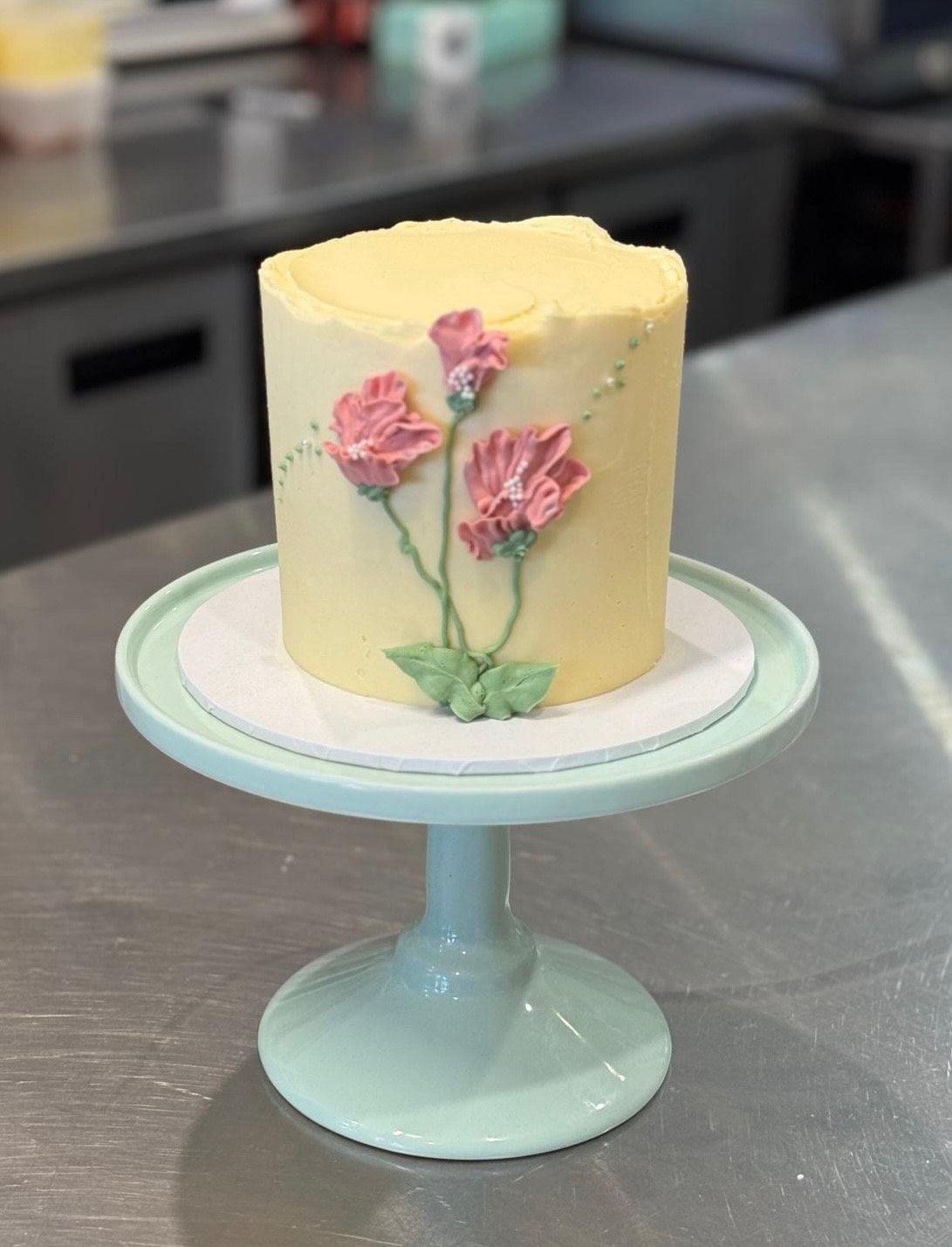 A round yellow cake decorated with pink flowers and green leaves, displayed on a light green cake stand.