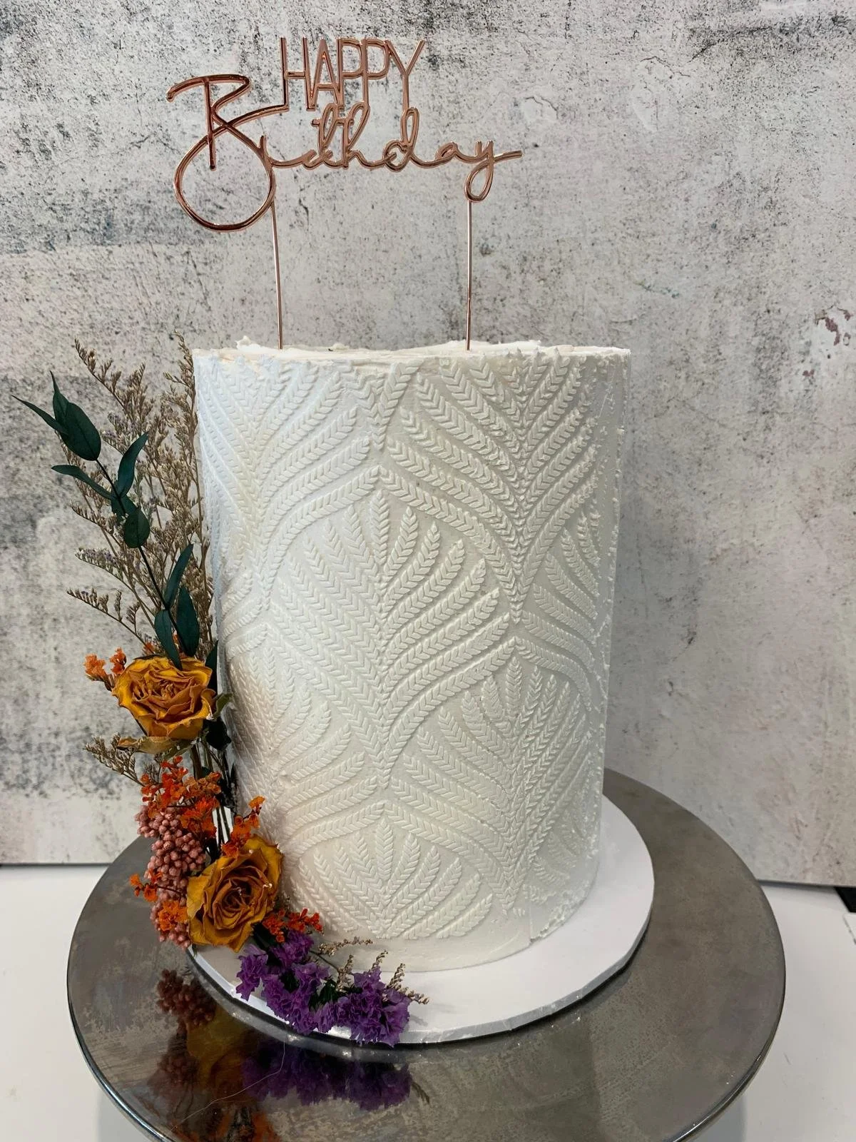 A white birthday cake with intricate textured leafy patterns, decorated with dried flowers and greenery on the side, topped with a copper-colored "Happy Birthday" cake topper, placed on a reflective silver cake stand against a light textured backgrou