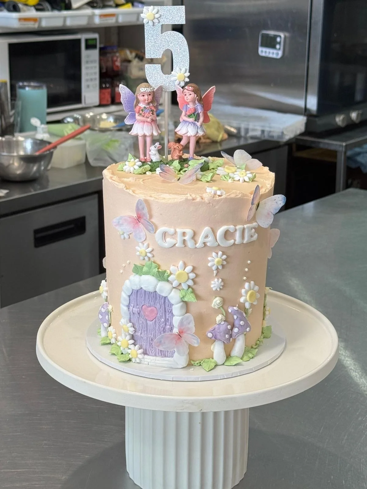 A pastel pink birthday cake decorated with edible flowers, butterflies, and fairy figurines on top, with a large number five cake topper. The cake has 