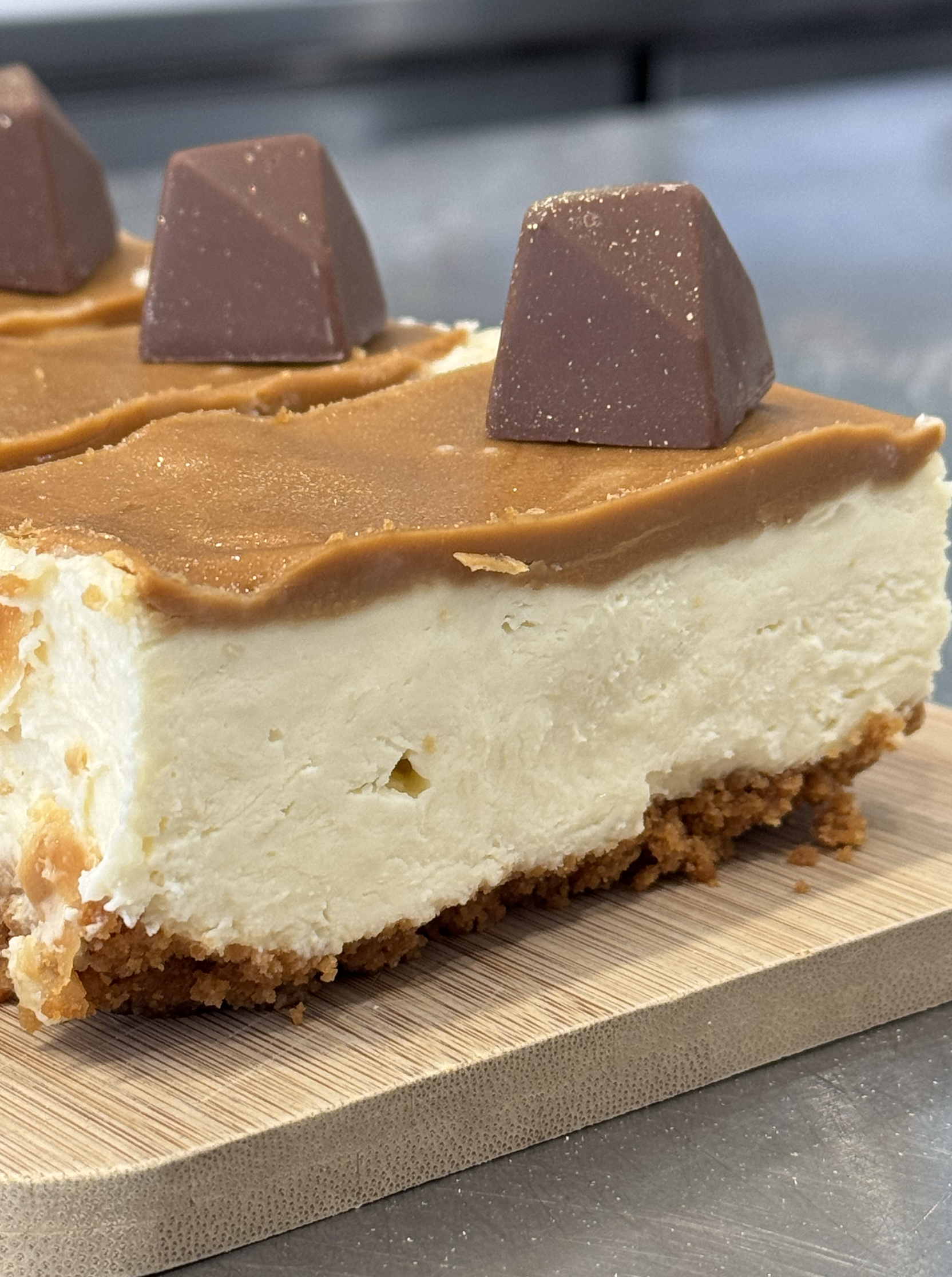 Close-up of a cheesecake topped with caramel and chocolate pieces, sitting on a wooden board.