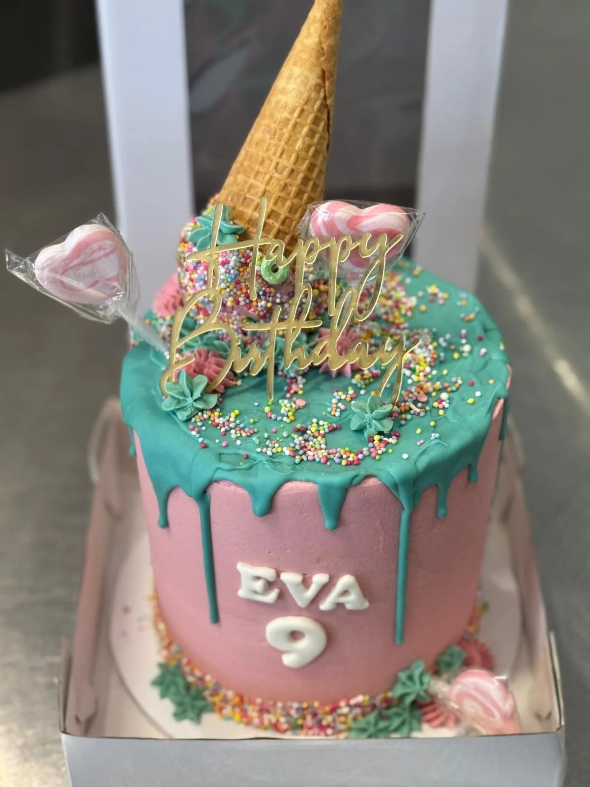 A pink birthday cake with turquoise drip icing, decorated with sprinkles and small frosting flowers. It has a golden 'Happy Birthday' topper, an inverted ice cream cone, and wrapped pink and white heart-shaped candies. The cake has the name 'EVA' and