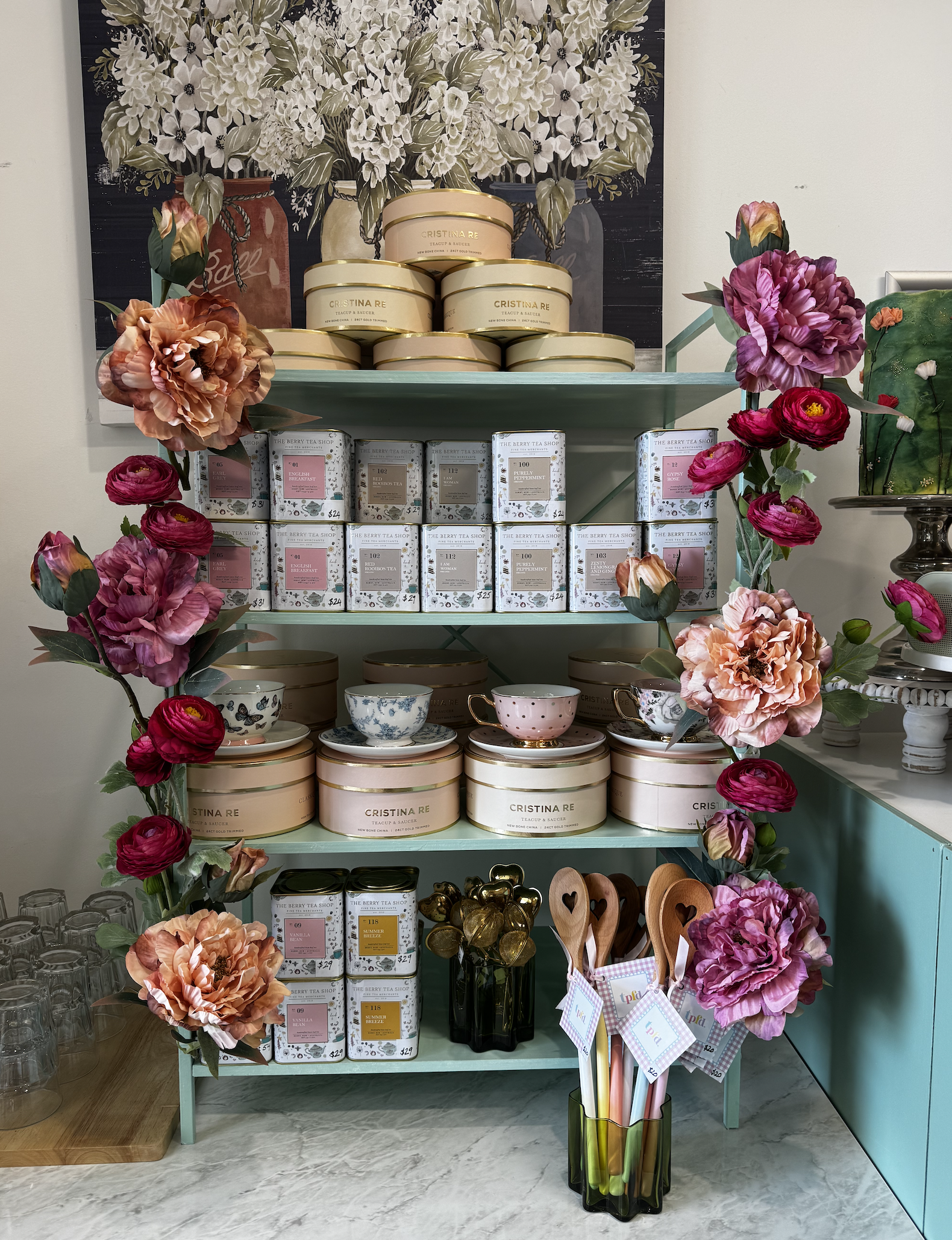 Display of tea tins, floral arrangements, and kitchen utensils on a mint green shelf with a floral artwork in the background.