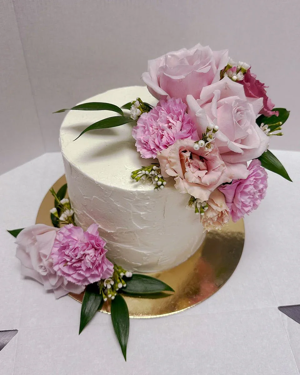 White cake decorated with pink roses, pink carnations, and green leaves.