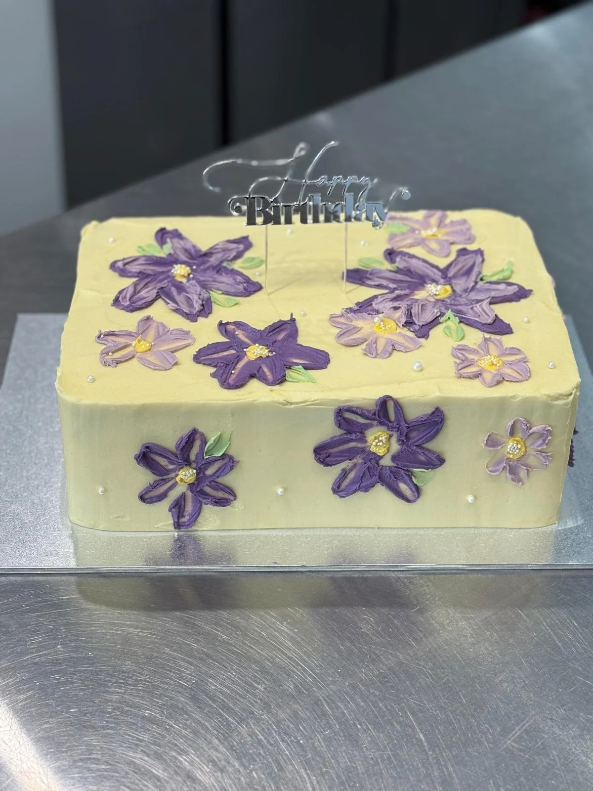 A rectangular birthday cake decorated with purple and lilac flower icing, small white pearl accents, and a silver 'Happy Birthday' topper.