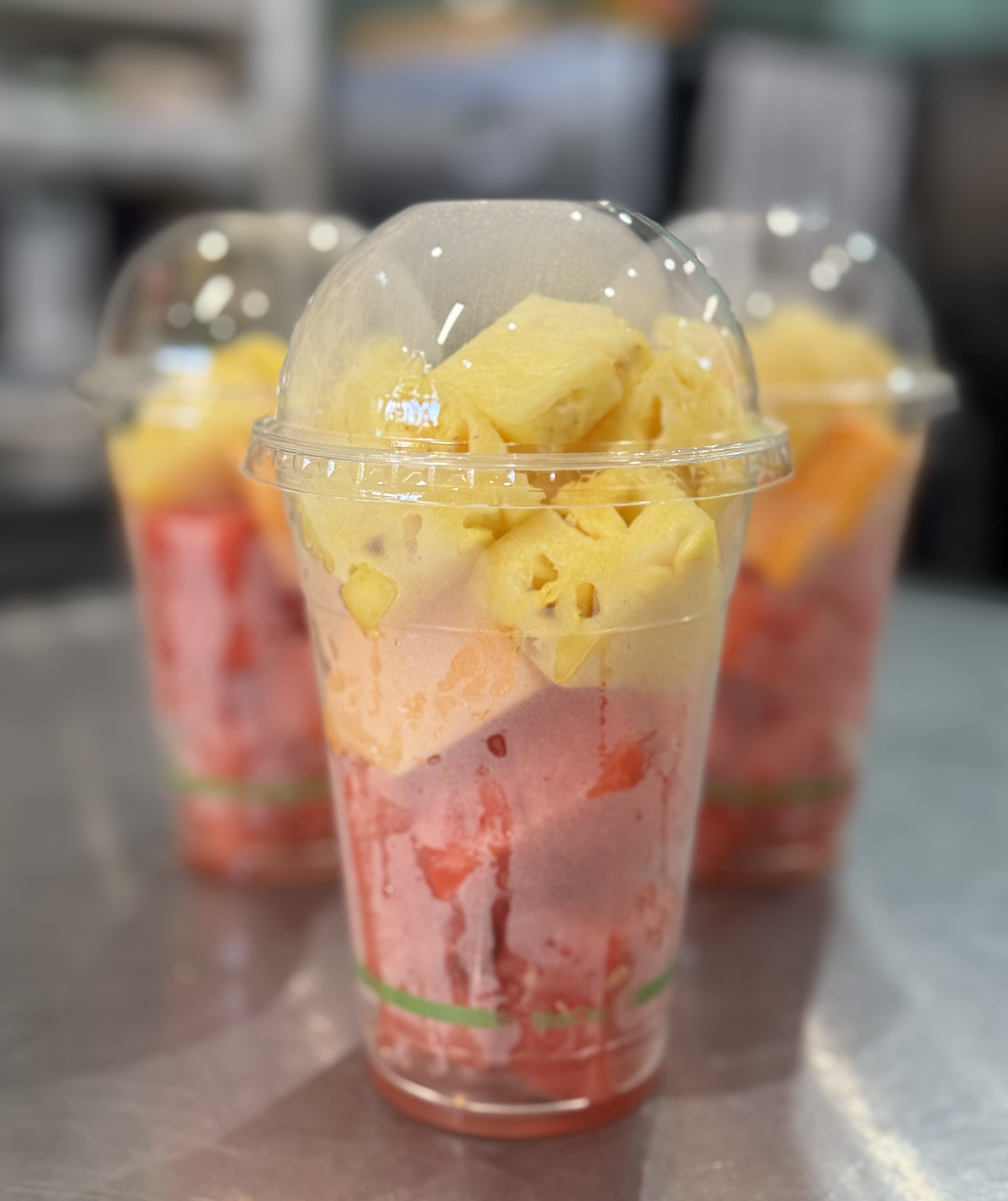 Three cups of fruit parfaits with layers of pink, yellow, and orange fruits, topped with pineapple chunks, in clear plastic cups with dome lids