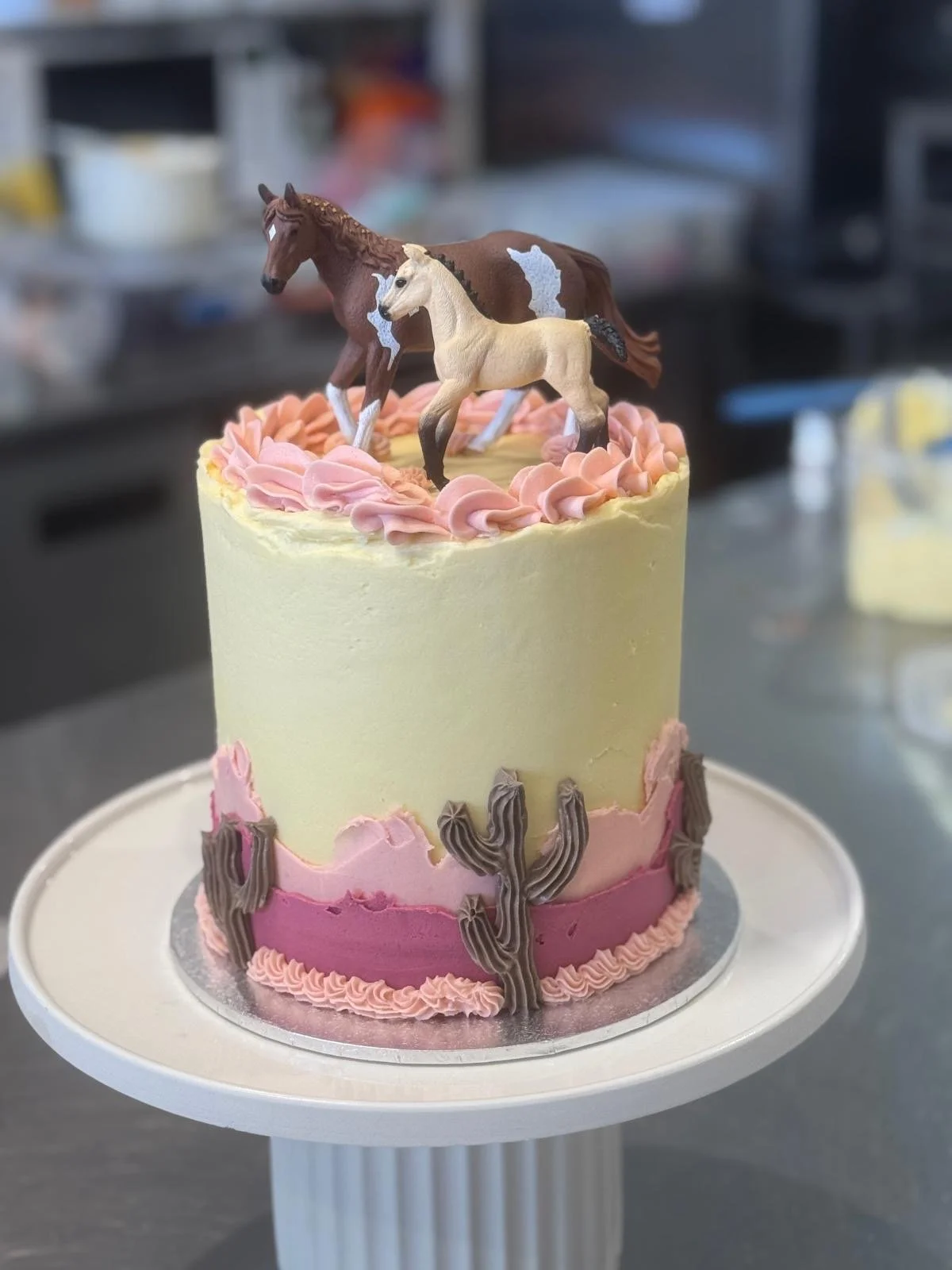 A colorful cake decorated with two toy horses on top, with pink and brown frosting and cactus-shaped decorations around the sides.