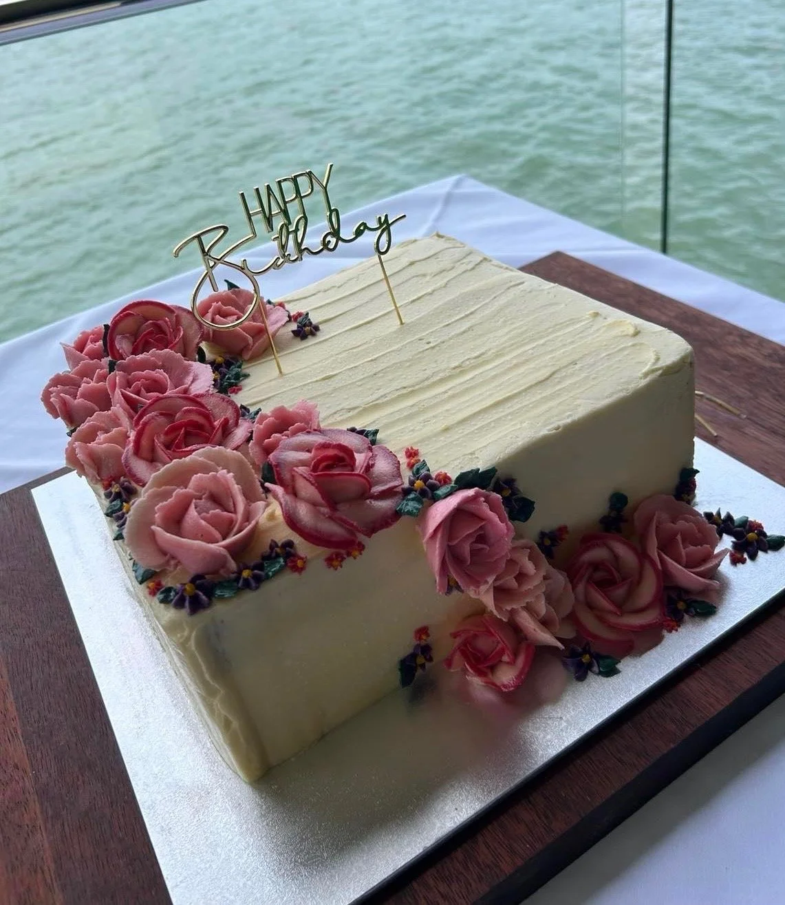 Decorated birthday cake with pink roses and a 'Happy Birthday' topper, placed on a table near water.