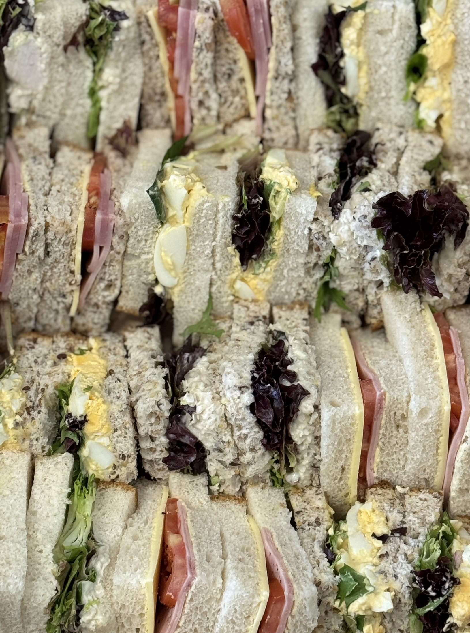 Close-up of assorted sandwich triangles with various fillings including lettuce, tomato, ham, egg salad, and cheese, arranged on a platter.