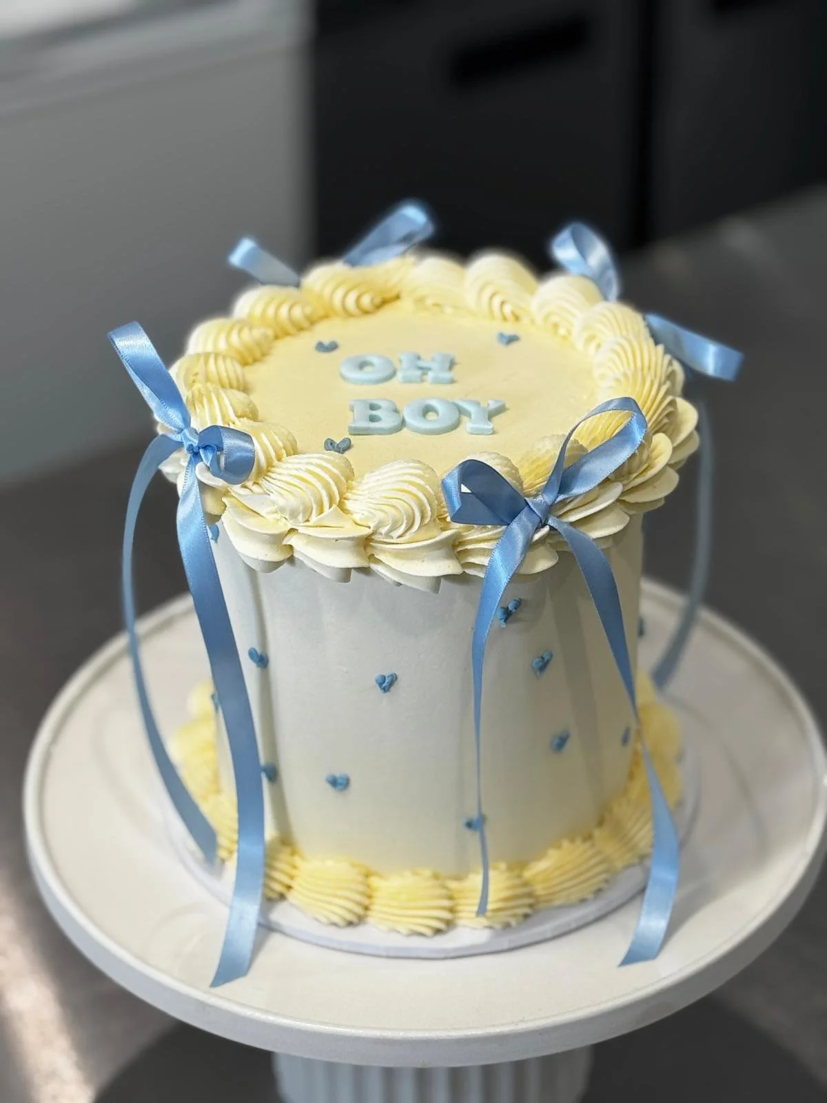 A cake with white and yellow frosting, decorated with blue ribbons, fondant letters spelling 'OH BOY', and small blue hearts.