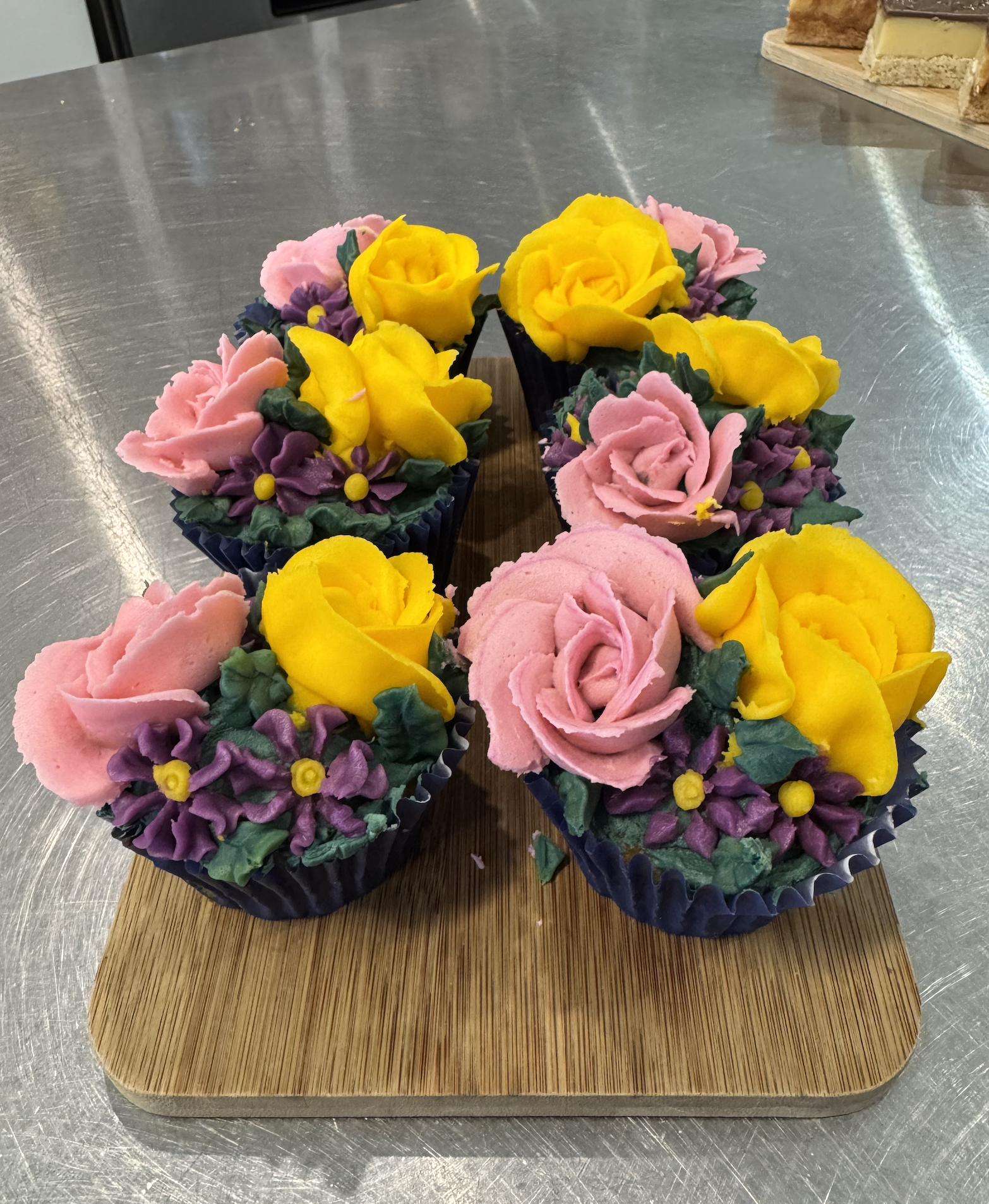 Cupcakes decorated with pink, yellow, and purple icing, arranged in black cupcake liners on a wooden tray.