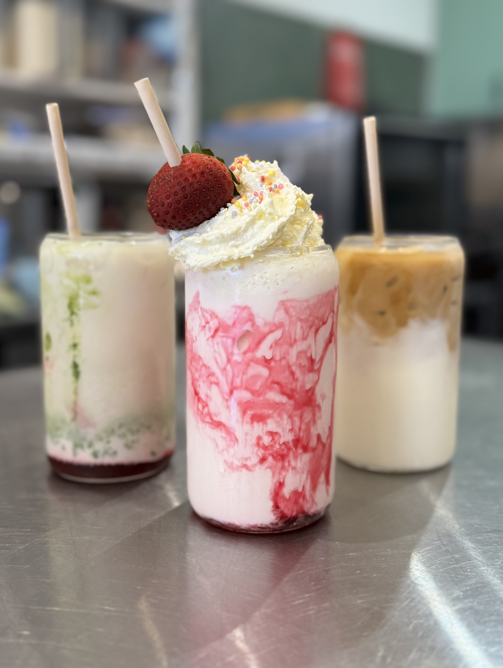 Three milkshakes topped with whipped cream, sprinkles, and strawberries, placed on a metal countertop in a café or restaurant.