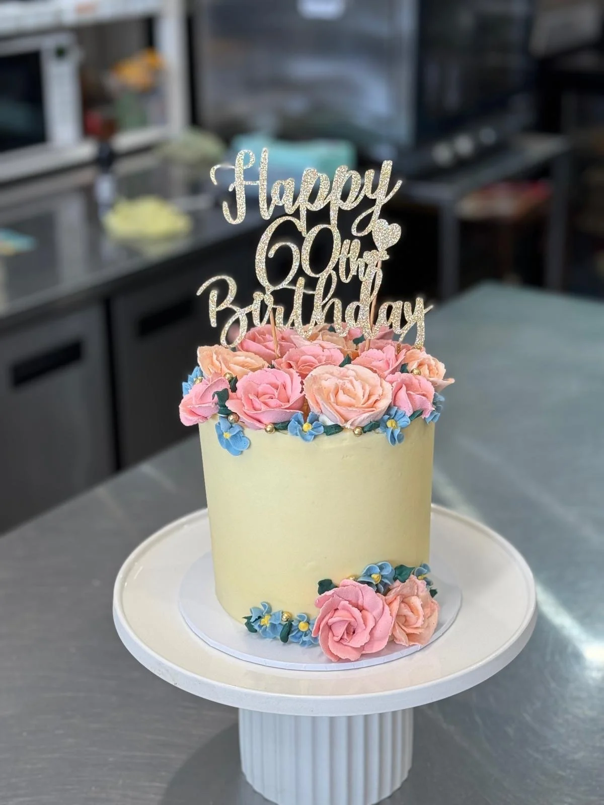 A birthday cake decorated with pink peonies and small blue flowers, topped with a glittery 'Happy 60th Birthday' cake topper, on a white cake stand in a kitchen.