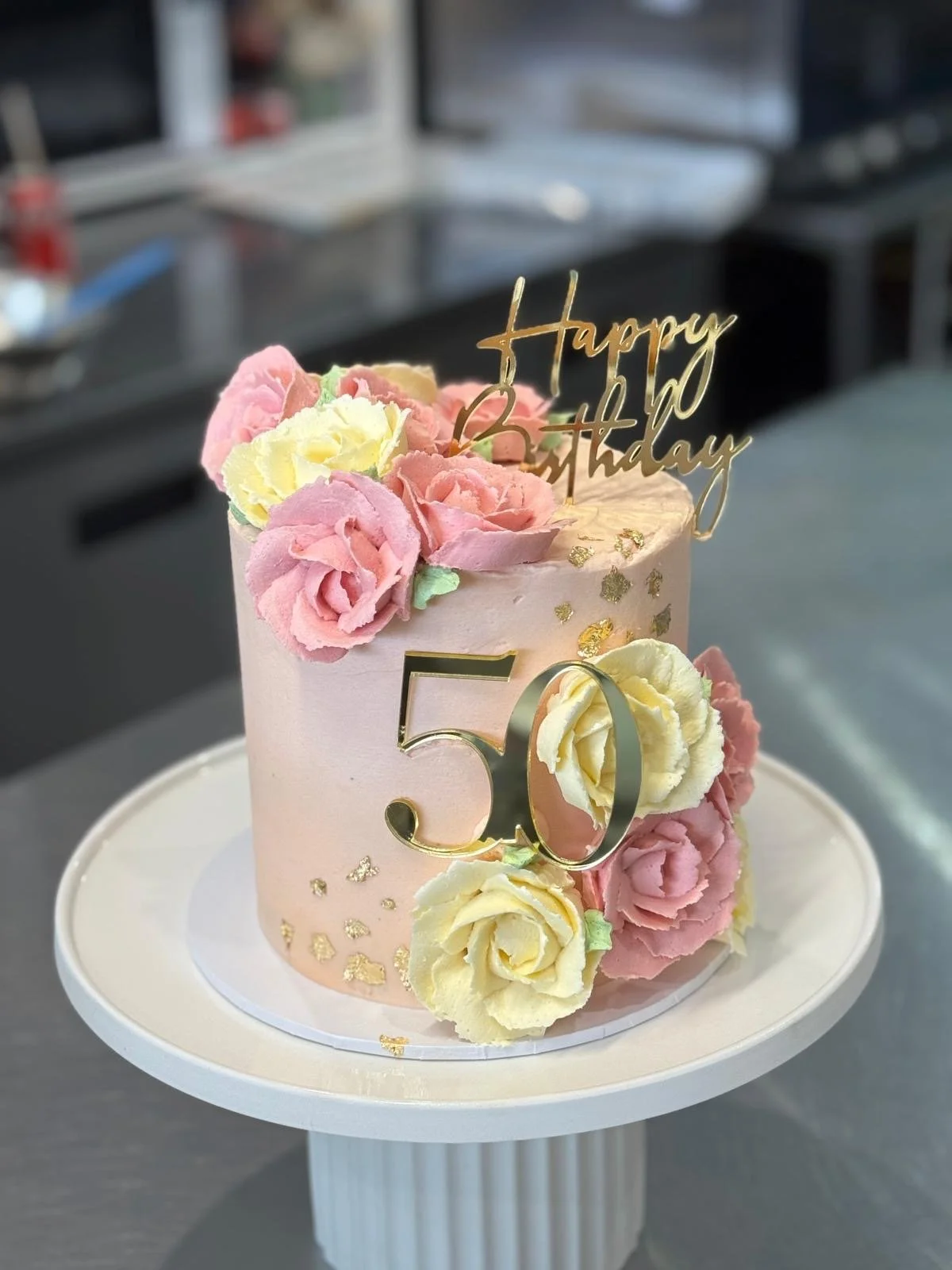 A pink birthday cake decorated with pink and yellow buttercream flowers, a gold 'Happy Birthday' topper, and a gold '50' sign with gold confetti accents, all on a white cake stand.