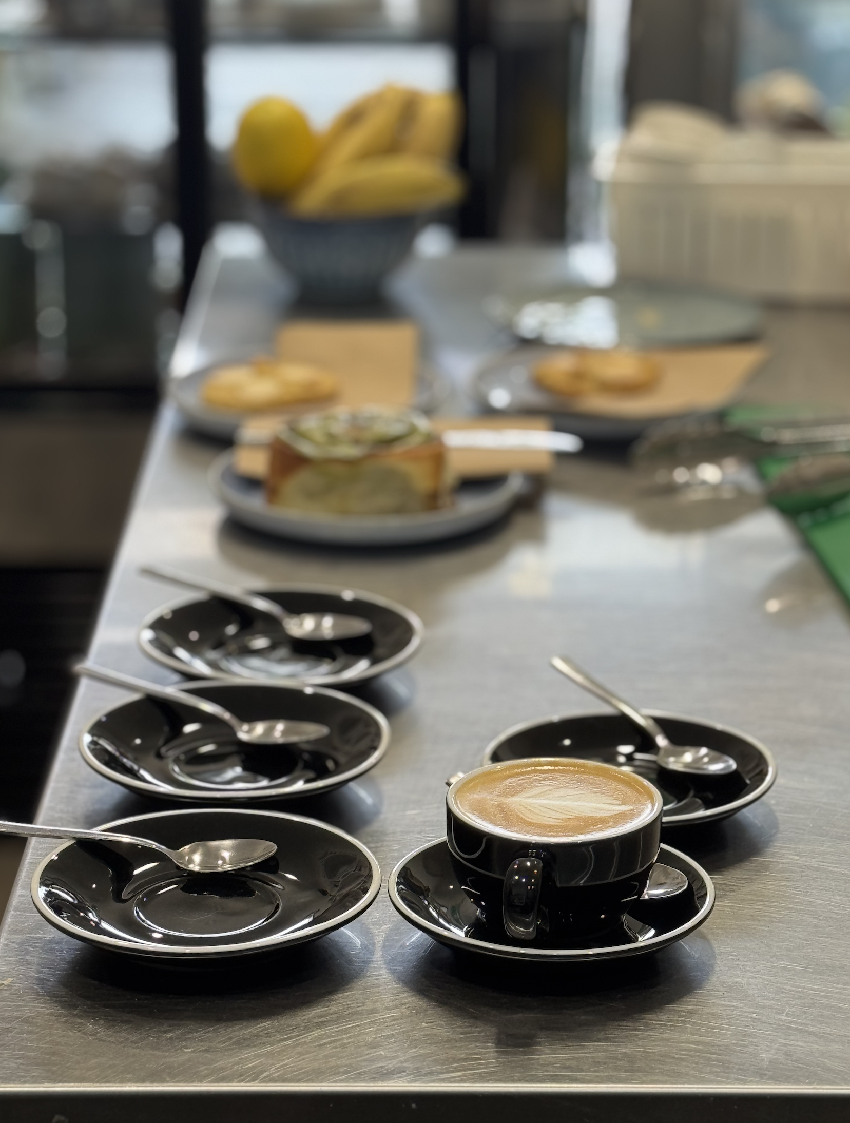 Several black coffee cups with saucers and teaspoons are arranged on a metal table, with a tray of pastries and cups of coffee in the background.
