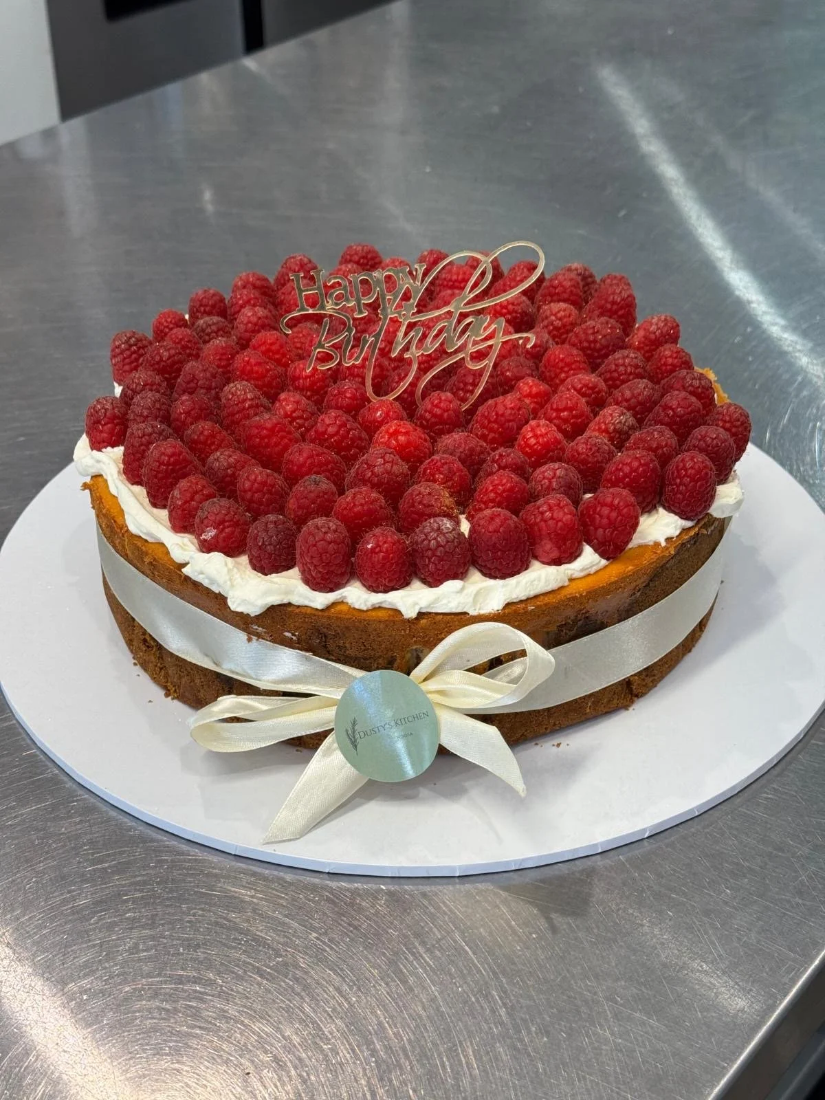Raspberry-topped birthday cake with a 'Happy Birthday' topper, white ribbon around the base, and a logo tag, on a white platter.