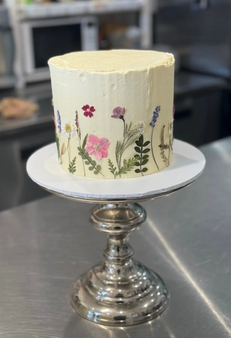 A cake decorated with dried pressed flowers and greenery, placed on a silver cake stand in a professional kitchen.