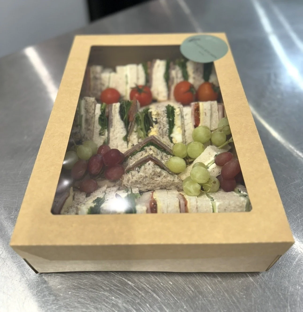 A cardboard box with a clear plastic top containing assorted small sandwiches, grapes, and cherry tomatoes on a metal surface.