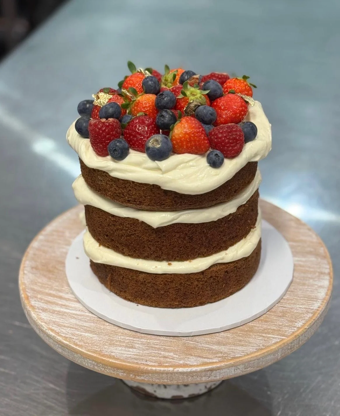 Layered chocolate cake with cream cheese frosting and topped with fresh strawberries, blueberries, raspberries, and small gold leaf accents, on a wooden cake stand.