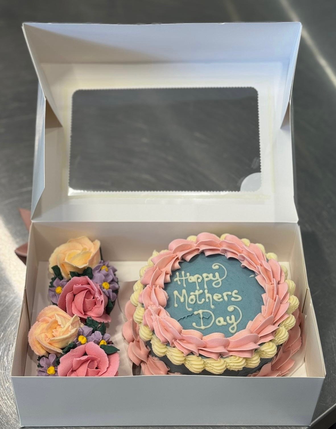 A Mother’s Day cake in a white box with pink and cream rosette decorations and a message that says 'Happy Mothers Day' written on a blue fondant circle.