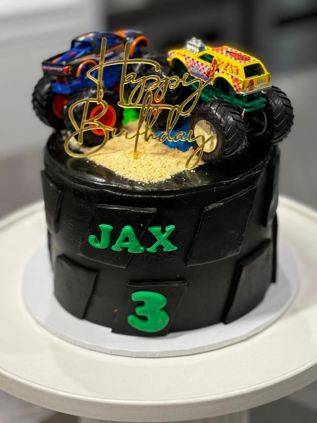 A birthday cake with a black tire design and green lettering that reads JAX and 3. On top, there are two toy monster trucks and a gold 'Happy Birthday' cake topper. The cake is decorated with yellow sprinkles and is placed on a white circular cake bo