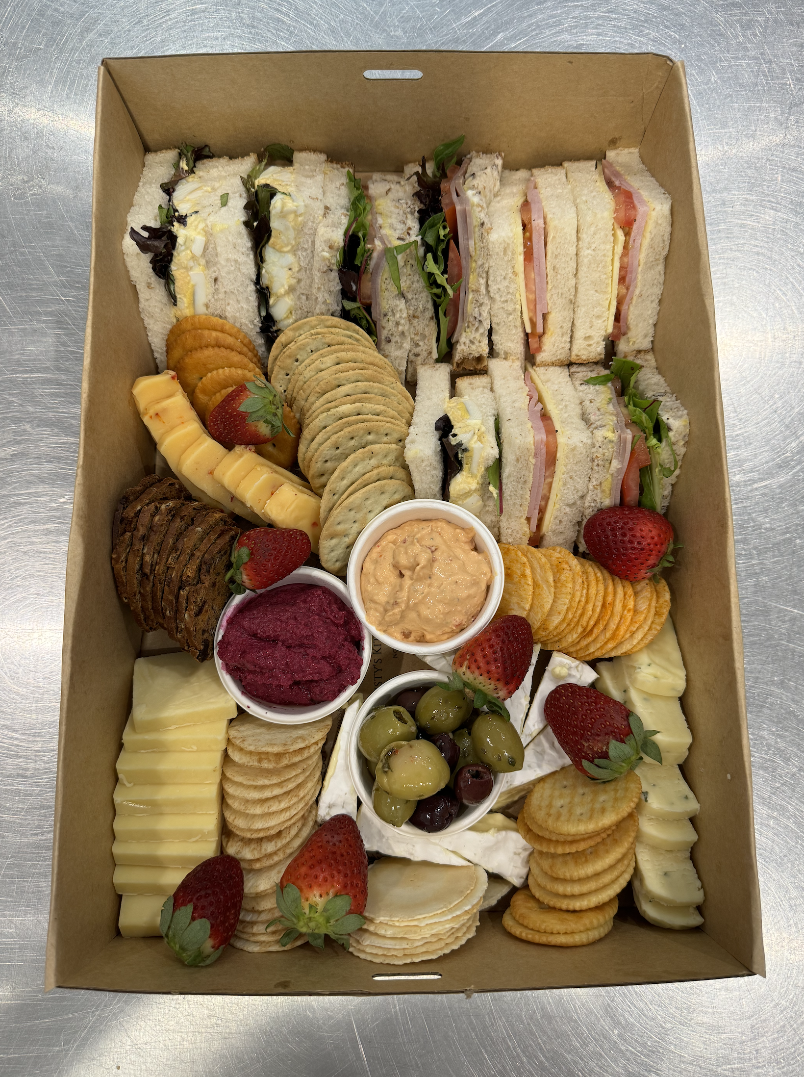 A box filled with assorted sandwiches, cookies, strawberries, grapes, cheese, crackers, and dipping sauces, arranged on a metal surface.