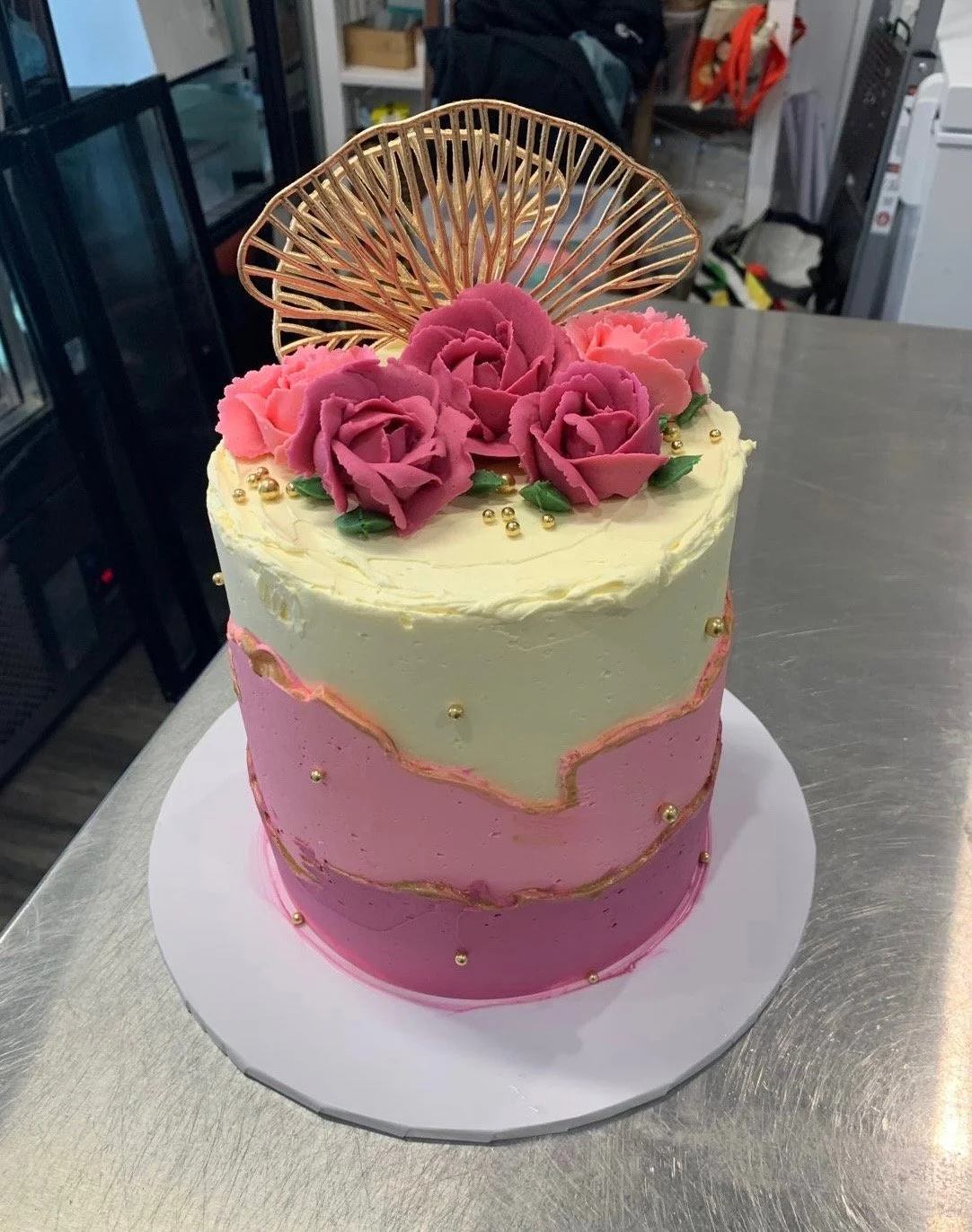 A two-tier pink and white frosted cake decorated with pink and purple sugar roses, green leaves, gold beads, and a gold decorative topper, sitting on a white cake board.