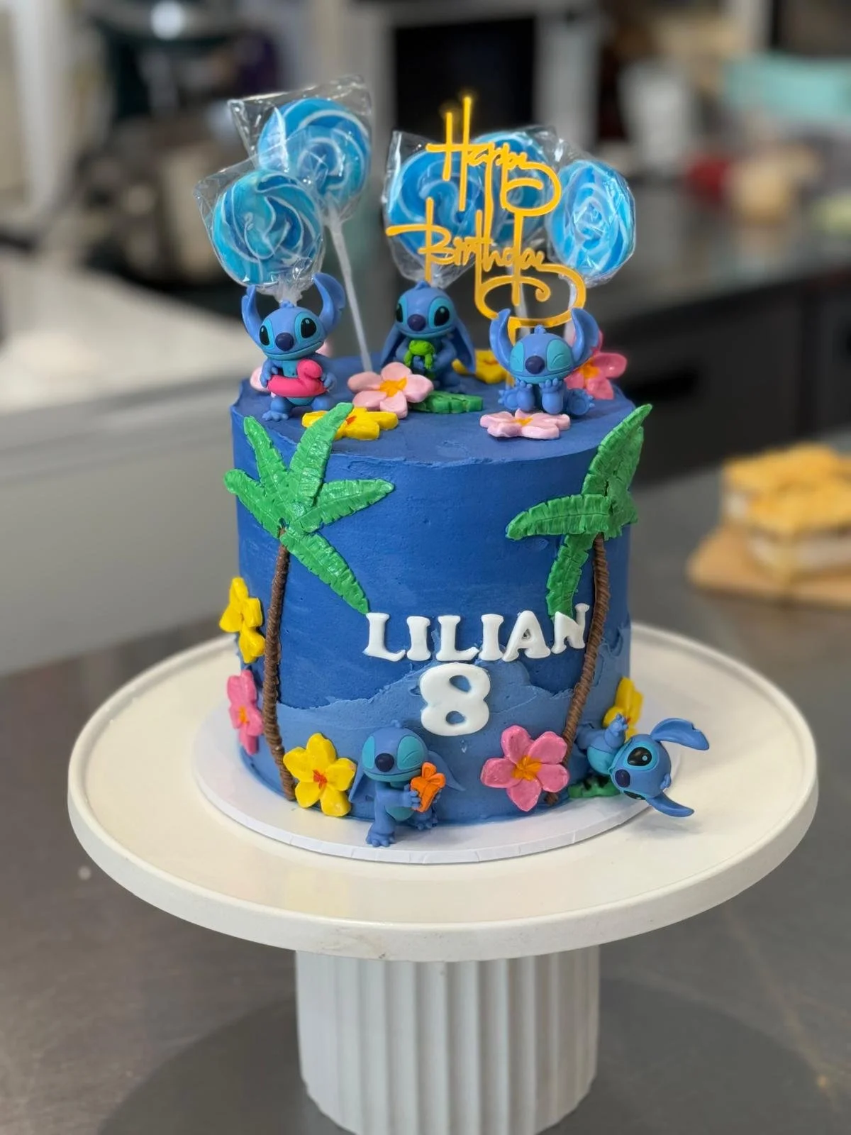 Blue birthday cake decorated with Stitch figures, pink and yellow flowers, palm trees, and a yellow "Happy Birthday" topper. The cake has the name Lillian and the number 8, indicating an 8th birthday.