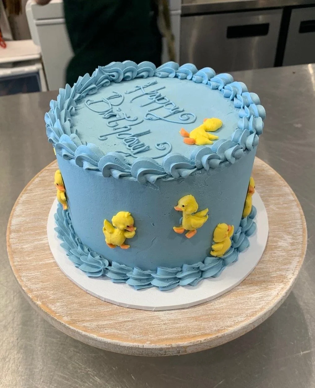 Blue birthday cake decorated with yellow duck figurines, piped blue icing border, and the words "Happy Birthday" written on top.