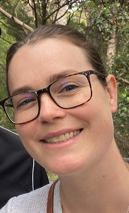Close-up of a smiling woman wearing glasses outdoors with trees in the background.