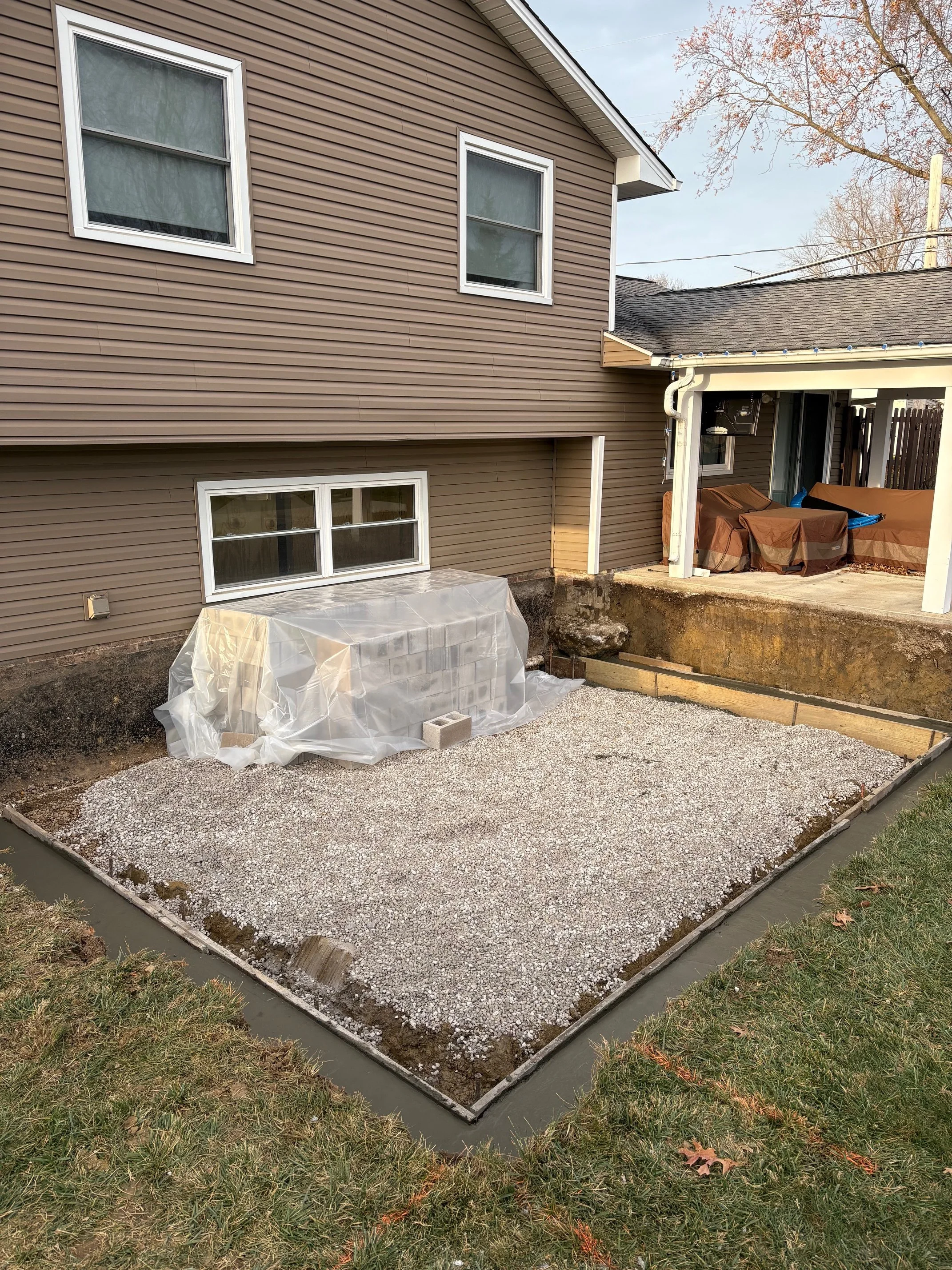 Backyard construction site with gravel and a covered stack of bricks next to a house with brown siding and white-framed windows.